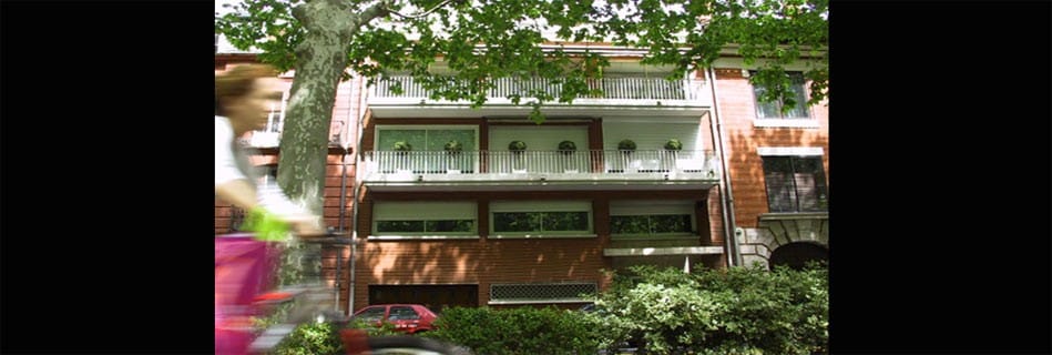 Façade sur rue de l'immeuble en brique toulousaine, balcons à garde-corps blancs et platanes, une cycliste passant devant
