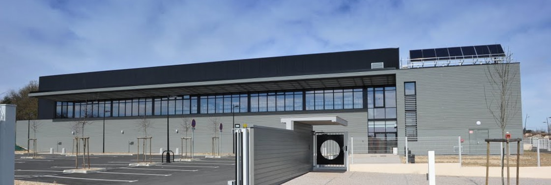 Façade du gymnase en bardage métallique gris avec longue verrière et panneaux solaires en toiture, parvis et parking plantés