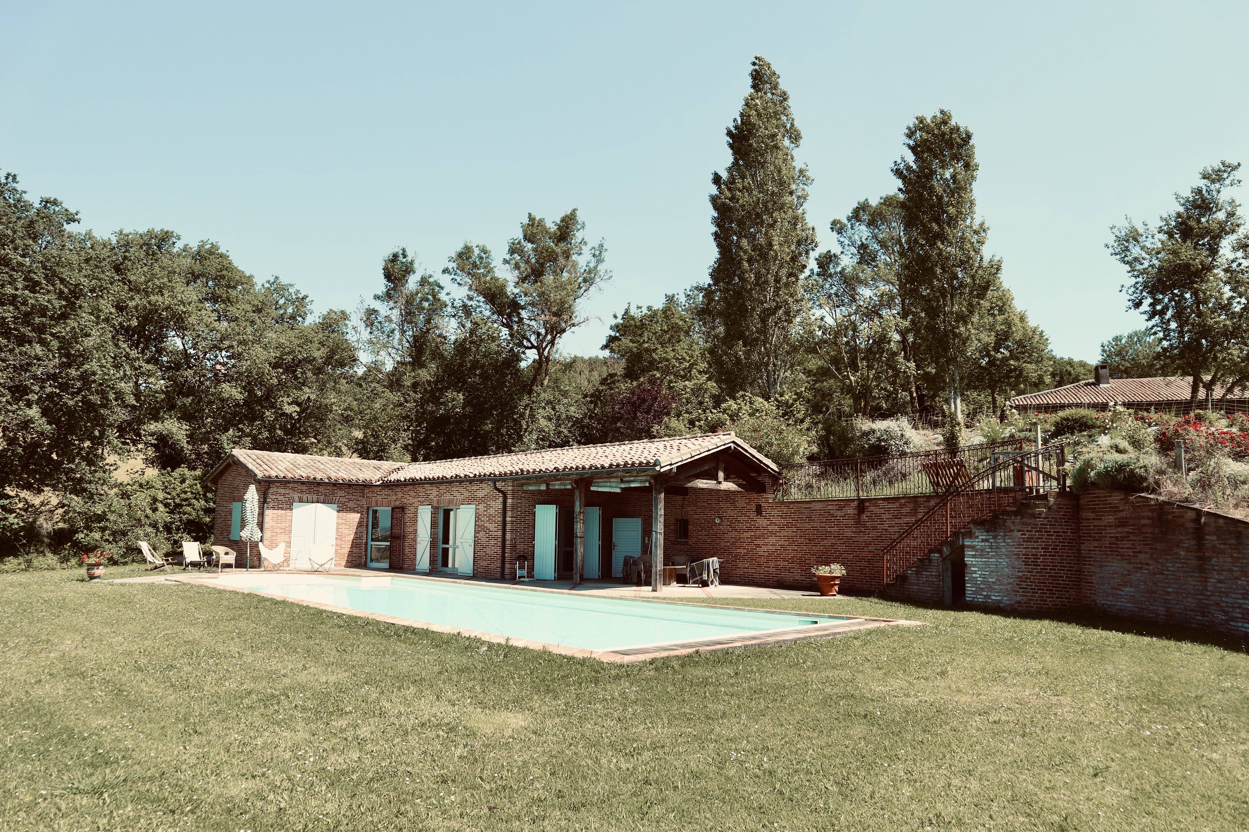 Maison en brique bâtie dans la pente avec pool house aux volets bleus, piscine, mur de soutènement en brique et escalier métallique montant vers le jardin