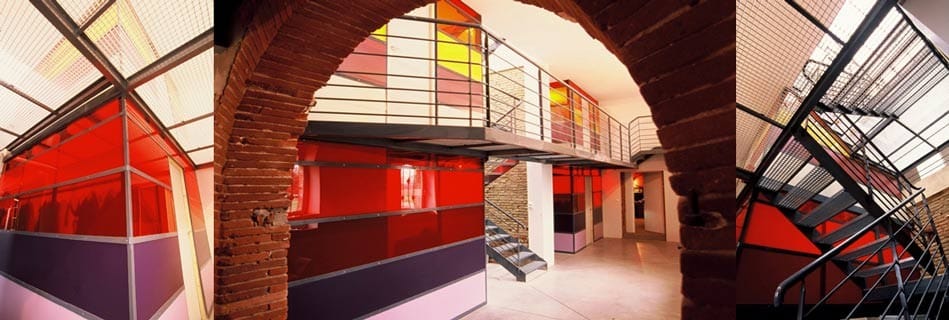 Les totems : blocs de verre coloré rouge, orange et jaune reliés par une passerelle en caillebotis métallique sous les arches de brique